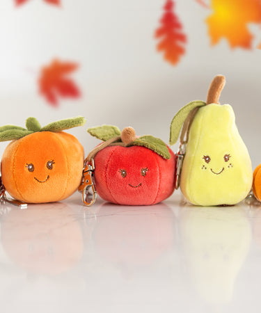 A collection of plush key rings aligned in a row including a strawberry, orange, red apple, green pear, and pumpkin with leaves falling from above.