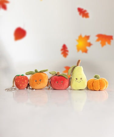 A collection of plush key rings aligned in a row including a strawberry, orange, red apple, green pear, and pumpkin with leaves falling from above.