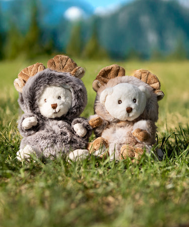 Two plush bears dressed in moose outfits sit in a patch of grass, with tall, majestic mountains rising in the background under a clear blue sky