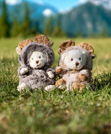 Two plush bears dressed in moose outfits sit in a patch of grass, with tall, majestic mountains rising in the background under a clear blue sky